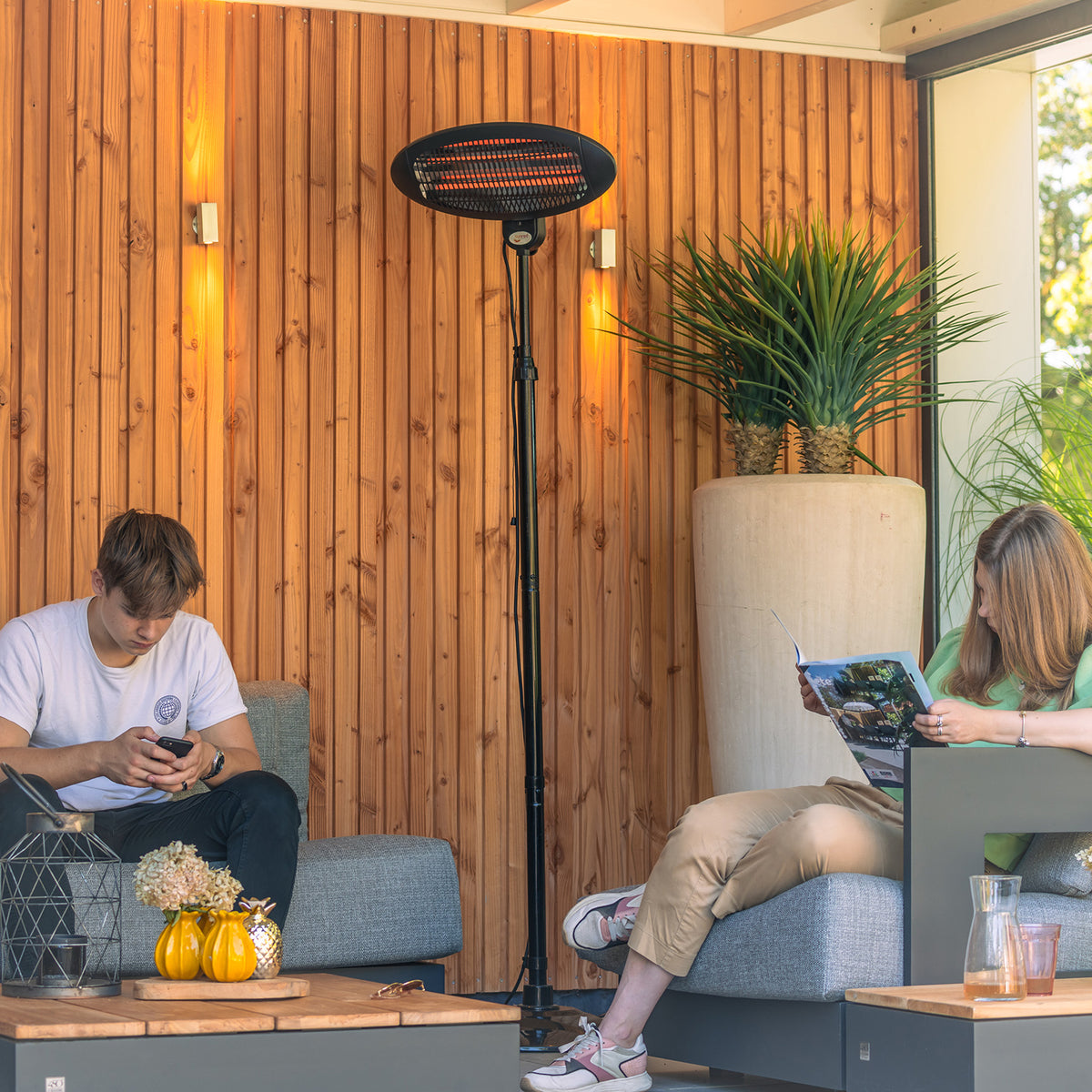A black floor-mounted patio heater stands between two people sitting on gray outdoor lounge chairs in a modern wooden patio setting. The heater has an oval heated element at the top and is shown in use in a lifestyle context.