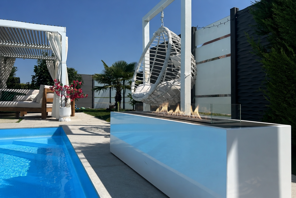 White freestanding bio fireplace used outdoors beside a pool, lit with a soft natural flame, hanging egg chair and palm trees